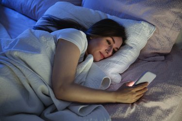 A smiling person laying in bed looking at their cellphone