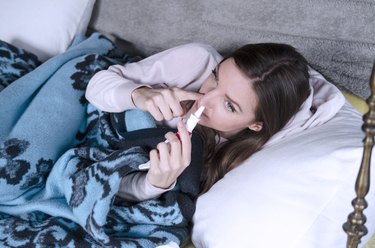 a person with long brown hair lying on the couch and using nasal spray because they have a stuffy nose at night