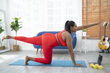 Person wearing red sports bra and leggings doing a strength-building Pilates workout on exercise mat at home.
