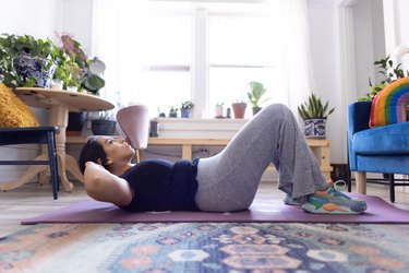 A person in grey pants and a dark blue shirt doing crunches on a purple yoga mat in their apartment, as a way to reduce stomach creases