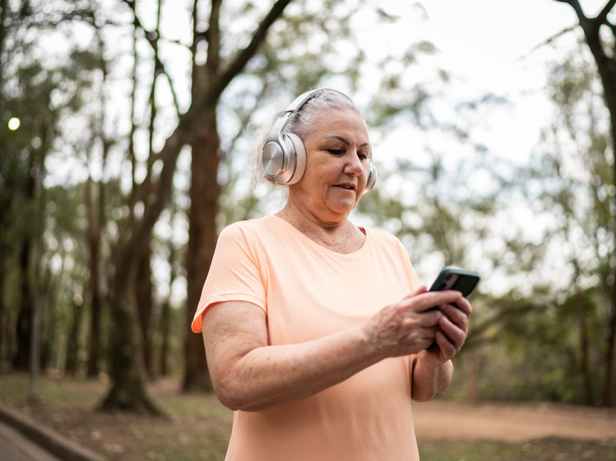 an older adult uses a mobile phone to play a workout playlist on silver headphone in a park as an example of workout motivation tips from fitness professionals