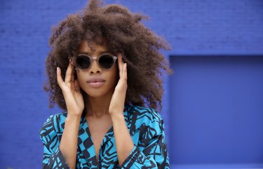 a young adult with natural hair wearing sunglasses in front of a blue wall