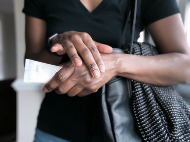 close view of a person wearing a black t-shirt holding a white lotion bottle and applying hand cream to their dry skin