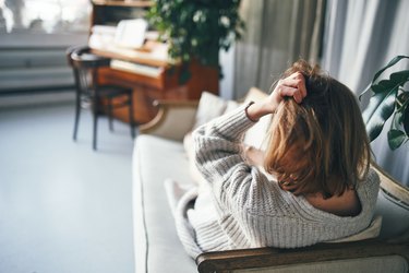 a person with shoulder-length light brown hair wearing a grey sweater lying on a couch with their back to the camera and holding their head because they have a headache after dental work
