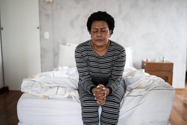 Person in black and white striped pajamas sitting on the edge of the bed with hands clasped and an uncomfortable facial expression