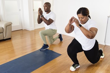 Two people performing body-weight lunges in their living room to demonstrate a lower-body workout with no equipment.