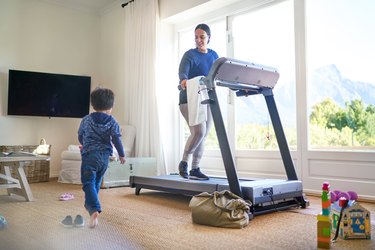 Exerciser walking on treadmill in living room while child walks by.