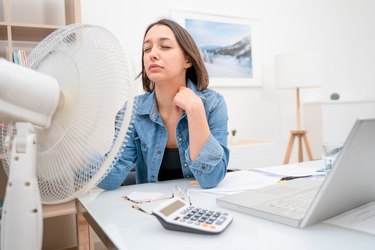 a person with shoulder-length brown hair sitting at a desk in front of a fan to cool down because they're always hot