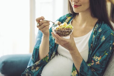 A smiling pregnant person wearing a green kimono and eating a bowl of granola topped with fruit