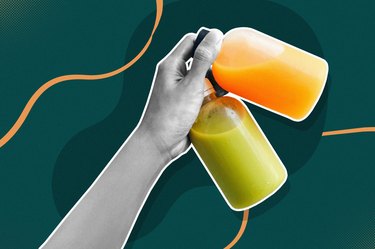 close up of hand holding glass bottles of green and orange juice for a juice cleanse on a dark green background