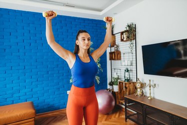 woman wearing bright blue tank and orange leggings doing a shoulder dropset dumbbell workout at home