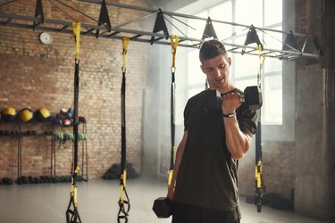 man doing biceps curls with dumbbells during upper-body workout while standing against of brick wall at gym